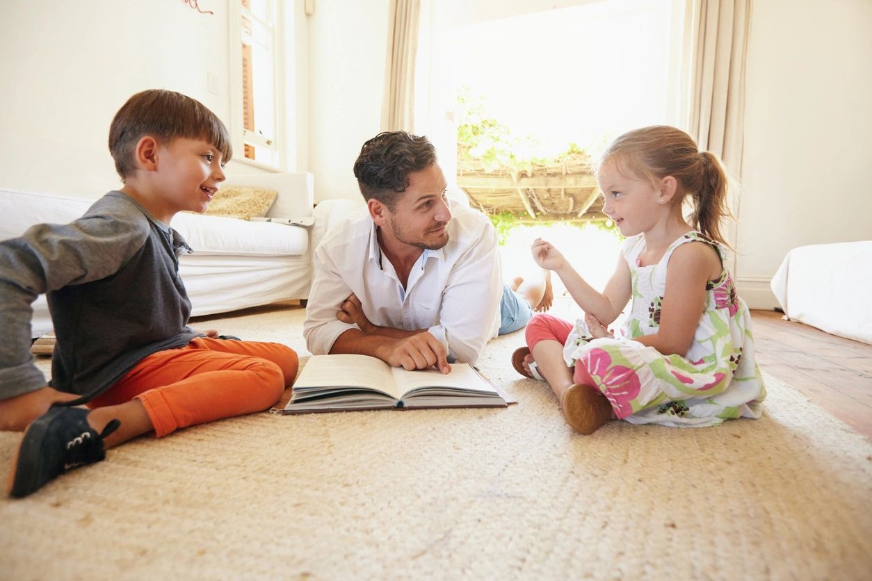 Family with book sitting on floor with little girl talking to his father and brother. Father with his little boy and girl at home.